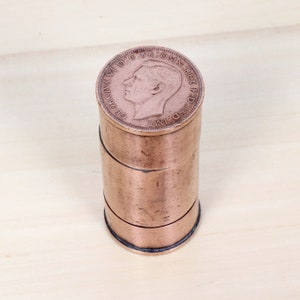 May include: A copper-colored cylindrical container with a coin on top. The coin is a British penny with the image of King George VI on the obverse.