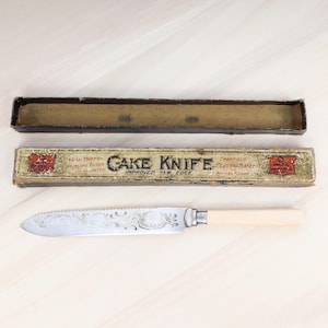 May include: An antique cake knife with a decorative blade and a cream-colored handle, displayed with its original box. The box is labeled "CAKE KNIFE" and features text indicating it is sterling silver.