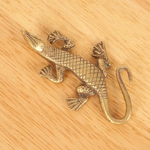 Könnte beinhalten: Eine goldfarbene Metallfigur eines Leguan mit einer strukturierten Oberfläche und einem eingerollten Schwanz. Der Leguan ist in einer Pose mit ausgestreckten Beinen und leicht nach oben geneigtem Kopf dargestellt.