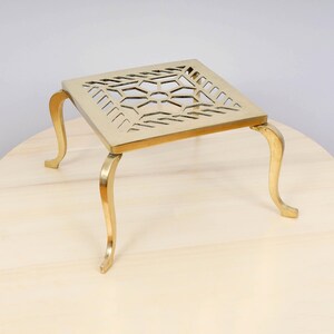 May include: A gold metal decorative stand with a square top and intricate cut-out design. The stand has four legs with a curved design.
