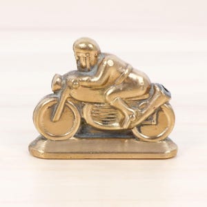 May include: A solid brass figurine depicting a motorcyclist in a racing crouch on a motorcycle. The metallic gold-toned object features a rider wearing a helmet, leaning forward over the fuel tank, mounted on a flat, rectangular base.