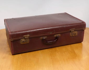 May include: A vintage burgundy suitcase with a textured surface. The rectangular case has brass-colored latches and a dark brown handle. The corners are reinforced with stitching, and the case appears to be made of a durable material.