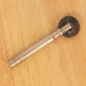 May include: A silver metal tool with a circular saw blade on one end and a handle on the other. The tool is lying on a wooden surface.
