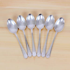 May include: Six silver spoons arranged on a light wood surface. Each spoon has a rounded bowl and a long, slender handle. The handles have a decorative design near the base and are engraved with the letter "M".