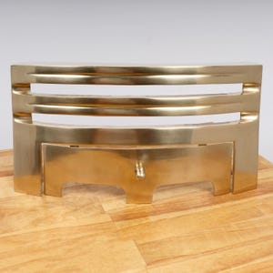 Könnte beinhalten: Ein Kaminfeuergitter aus poliertem Messing mit geschwungenem Frontdesign, drei horizontalen Lamellen und einer massiven unteren Platte. Die Metallstruktur verfügt über einen kleinen, mittig platzierten kugelförmigen Knopf im unteren Bereich und weist durchgehend eine glatte, reflektierende Metalloberfläche auf.