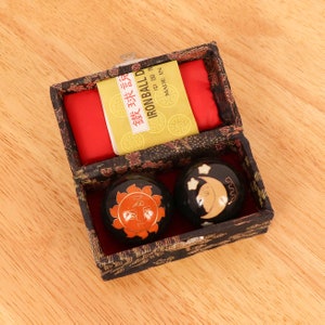 May include: A black box with a red lining and a yellow label that says "Ironball Double Balls" in Chinese and English. The box contains two black metal balls with gold designs of the sun and moon.