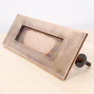 May include: A rectangular brass mail slot cover with a weathered, aged patina and visible surface scratches. The metal plate features a central recessed rectangular opening and includes a single threaded bolt with a metal washer on the back side.