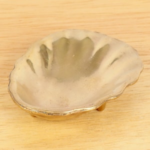 May include: A gold-coloured metal seashell dish with a scalloped edge and three small feet. The dish is empty and appears to be made of brass.