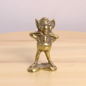 Peut inclure: Figurine en laiton représentant un gobelin souriant, les mains sur les hanches. Le gobelin a de grandes oreilles et un ventre rond. La figurine est posée sur une surface en bois.