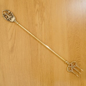 May include: A gold-toned fireplace poker with a decorative handle featuring a stylized figure. The poker has a long, slender shaft and a three-pronged tip.