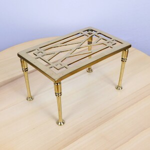 May include: A brass, geometric design hot pad with four legs. The hot pad is on a wooden surface.