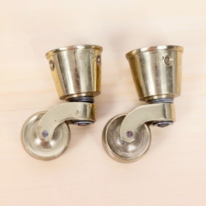 Könnte beinhalten: Zwei goldene Metall-Rollen mit Messing-Finish. Jedes Rad hat eine runde Oberseite und eine runde Basis. Die Rollen sind an einer Metallstange befestigt.