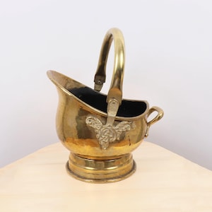 May include: A vintage brass coal scuttle with a curved handle and decorative embellishments. The scuttle has a rounded base and a small handle on the side. The interior is black. The scuttle is sitting on a light-colored wooden surface.