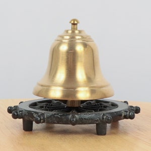 May include: A brass bell with a black cast iron base shaped like a ship's wheel. The bell is mounted on a central post and sits on top of the wheel.