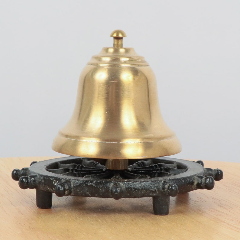 May include: A brass bell with a black cast iron base shaped like a ship's wheel. The bell is mounted on a central post and sits on top of the wheel.