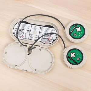 May include: An off-white retro gaming device with two circular controllers and a central unit. The controllers are round with green accents and directional pads. The central unit is open, revealing internal components and instructions.