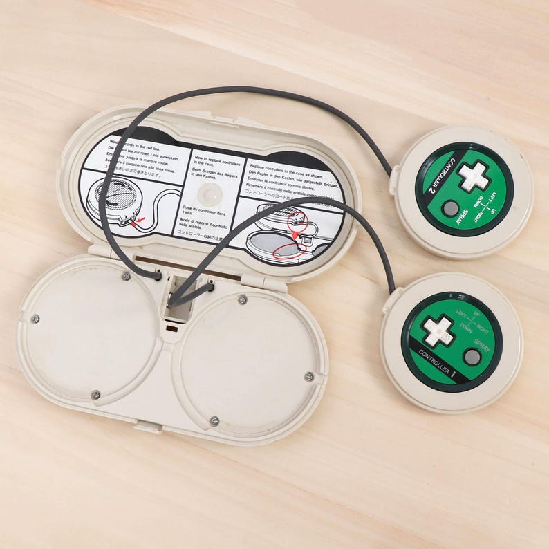 May include: An off-white retro gaming device with two circular controllers and a central unit. The controllers are round with green accents and directional pads. The central unit is open, revealing internal components and instructions.