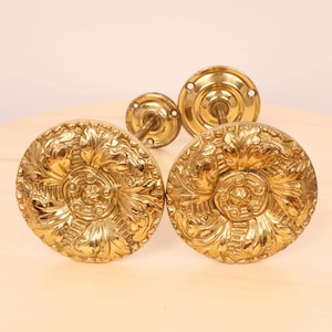 May include: Two gold-toned metal curtain tiebacks with ornate floral designs. The tiebacks have a round, flat base with a decorative floral pattern and a long, thin rod extending from the centre.