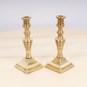Puede incluir: Un par de candelabros antiguos de latón con un diseño simple y elegante. Los candelabros tienen una base cuadrada y una columna estriada.