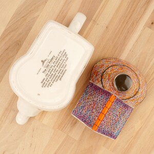 K&ouml;nnte beinhalten: Eine wei&szlig;e Teekanne mit einer bunten, mosaikartigen Vase. Die Teekanne tr&auml;gt ein Etikett mit der Aufschrift "The Old Pottery" und beschreibt den Prozess der T&ouml;pferherstellung.
