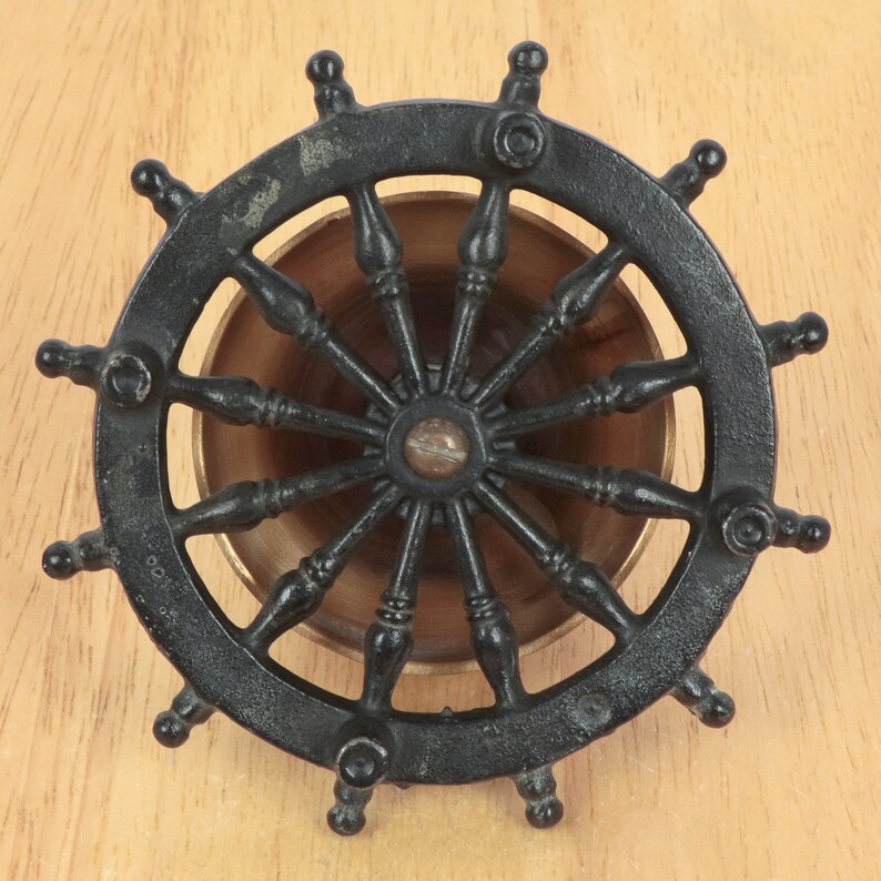May include: Black metal ship's wheel with a brass center and spokes. The wheel has a decorative design and is a nautical themed item.