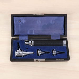 May include: A diagnostic otoscope set in a black rectangular case with a blue velvet interior. The set includes a black handle and three chrome-finished specula attachments. The text "MADE IN ENGLAND" is printed in white on the case exterior.