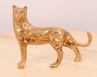 May include: A gold-colored metal figurine of a leopard with a spotted pattern. The figurine is standing on a light wood surface.
