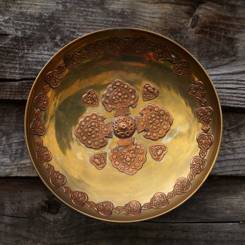 May include: A round, gold-colored metal serving dish with an intricate design of copper-colored hearts and floral patterns around the rim.