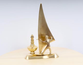 May include: A gold-toned metal sculpture of a person windsurfing. The sculpture is on a base with a small bottle.