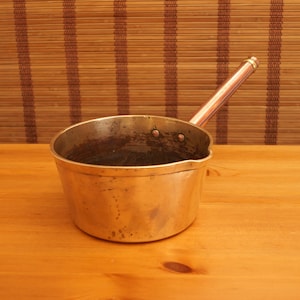 May include: A vintage brass saucepan with a copper handle. The saucepan has a worn, aged look and is slightly tarnished.