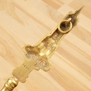 Peut inclure: Outil de chemin&eacute;e en m&eacute;tal dor&eacute; avec un motif d&eacute;coratif repr&eacute;sentant un b&acirc;timent. L'outil a un anneau sur le dessus et le num&eacute;ro "16861" est grav&eacute; sur le c&ocirc;t&eacute;.