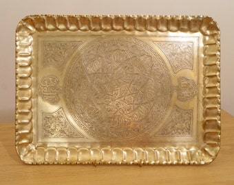 Puede incluir: Una bandeja de metal dorado con un diseño grabado intrincado. La bandeja tiene un borde festoneado y un diseño circular en el centro.