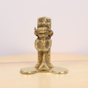 Peut inclure: Une figurine en laiton représentant un singe tenant une pelle, debout sur une base avec un motif décoratif. Le singe porte une couronne et a une expression espiègle.