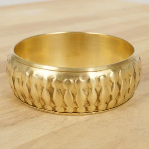 Könnte beinhalten: Ein goldfarbenes Metall-Armreif-Armband mit einem strukturierten, sich wiederholenden Muster aus kleinen, abgerundeten Formen.