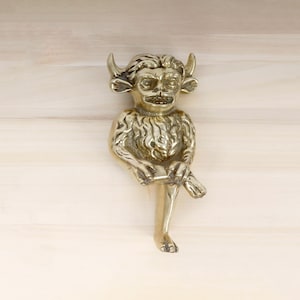 Peut inclure: Une sculpture en laiton de couleur dorée représentant une créature cornue, poilue et assise avec une expression espiègle. La sculpture est détaillée avec une fourrure texturée et un large sourire aux dents.