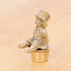 Può includere: Una statuetta in metallo dorato di un bambino con un cappello seduto su un vaso. La statuetta è un oggetto decorativo.