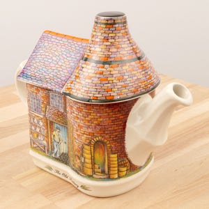 K&ouml;nnte beinhalten: Eine Keramik-Teekanne in Form eines Backsteingeb&auml;udes mit Schornstein. Die Teekanne ist mit einem Backsteinmuster und einer Szene eines Mannes, der in einer B&auml;ckerei arbeitet, verziert. Der Text "The Old Pottery" ist auf der Unterseite der Teekanne aufgedruckt.