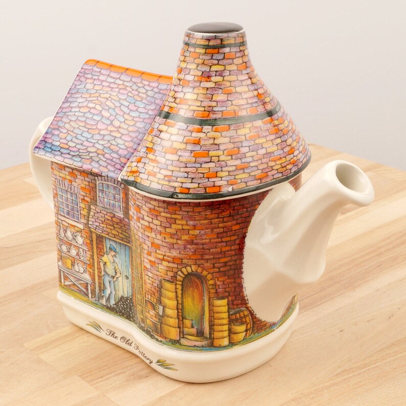 K&ouml;nnte beinhalten: Eine Keramik-Teekanne in Form eines Backsteingeb&auml;udes mit Schornstein. Die Teekanne ist mit einem Backsteinmuster und einer Szene eines Mannes, der in einer B&auml;ckerei arbeitet, verziert. Der Text "The Old Pottery" ist auf der Unterseite der Teekanne aufgedruckt.