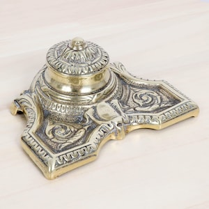 May include: A vintage brass inkwell with a detailed floral and scroll design. The inkwell has a hinged lid and sits on a decorative base. The brass has a warm, aged patina, adding to its antique charm. The inkwell is a classic desk accessory.
