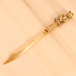 May include: A gold-toned letter opener with a detailed figurine of a seated figure at the handle.