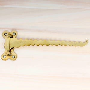 Puede incluir: Una bisagra de metal dorado con un borde decorativo festoneado. La bisagra está unida a una superficie de madera.