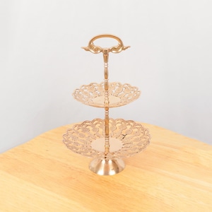 Puede incluir: Un soporte de servicio de latón de dos niveles con un diseño decorativo de filigrana. El soporte tiene un asa en el nivel superior y una base redonda.