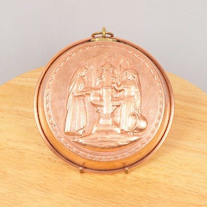 May include: Circular copper wall plaque featuring a relief of two figures in robes at a fountain. The copper has a warm, reddish-brown colour. A brass loop is at the top for hanging. The design is framed by a decorative border.