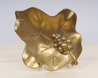 May include: A brass leaf-shaped dish with a raised design of grapes. The dish is a decorative piece with a vintage aesthetic.