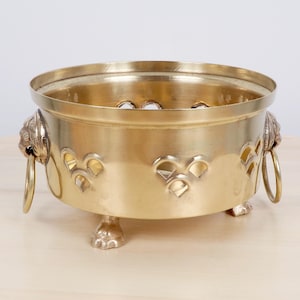 Può includere: Una ciotola in ottone con un design decorativo. La ciotola ha una forma circolare con un bordo largo ed è sostenuta da tre piedini a forma di zampa. Le maniglie a testa di leone sono attaccate ai lati. La ciotola ha ritagli a forma di cuore.