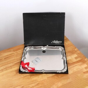 Op de afbeelding: Een rechthoekige zilverkleurige serveerschaal met sierlijke randen, gepresenteerd in een zwarte doos. De schaal heeft een reflecterend oppervlak en een label met een rood lint. De doos is gemarkeerd met "Ashberry Collection".