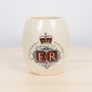 May include: Cream-colored ceramic mug with a commemorative design. The design includes a crown, the initials "ER" in a red and gold frame, and the text "Queen Elizabeth II Silver Jubilee 1977".