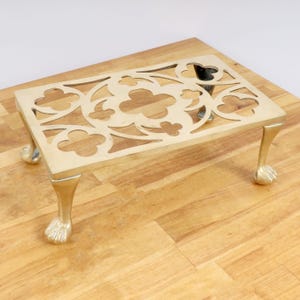 May include: A brass trivet with a decorative, intricate design and four paw-shaped legs. The trivet is on a wooden surface.