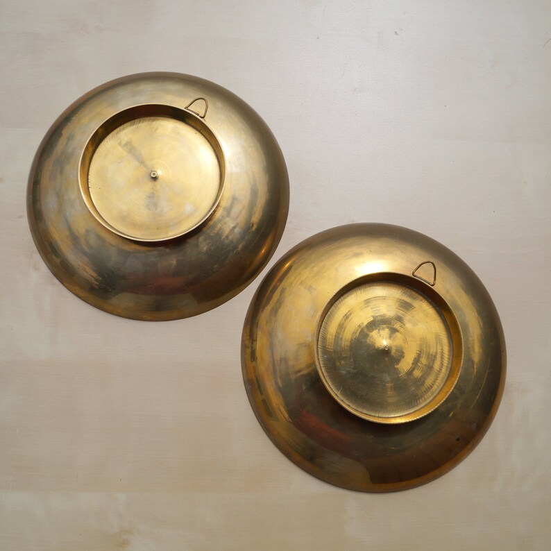 May include: Two round, brass wall plates with a decorative design. Each plate has a small, round handle on the back.