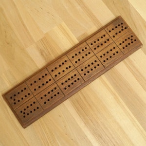 Può includere: Un tabellone da cribbage in legno con 12 file di buchi. Il tabellone è in legno marrone scuro e ha una finitura naturale.